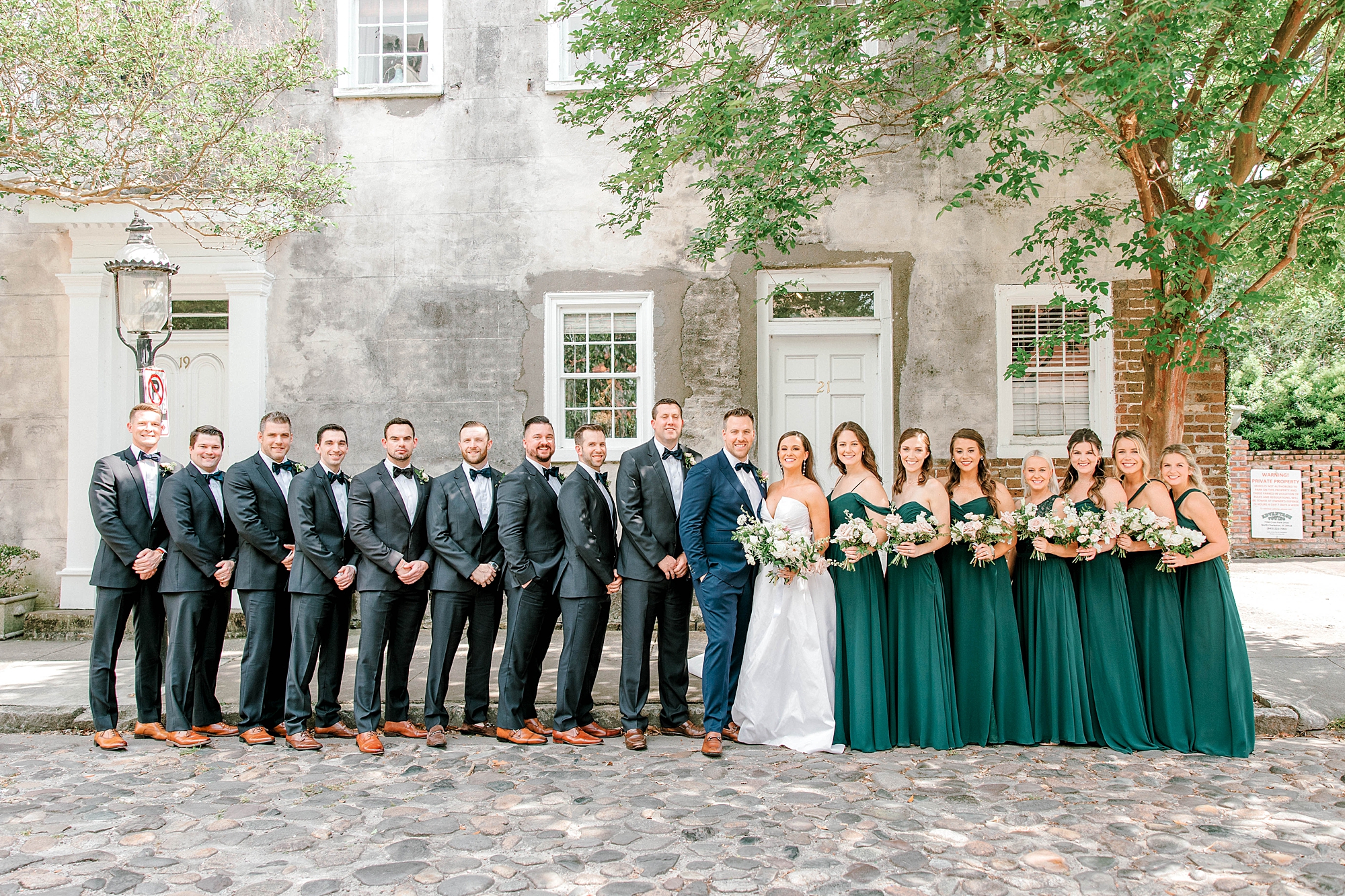 bride and groom stand with bridesmaids in green dresses and groomsmen in navy suits 