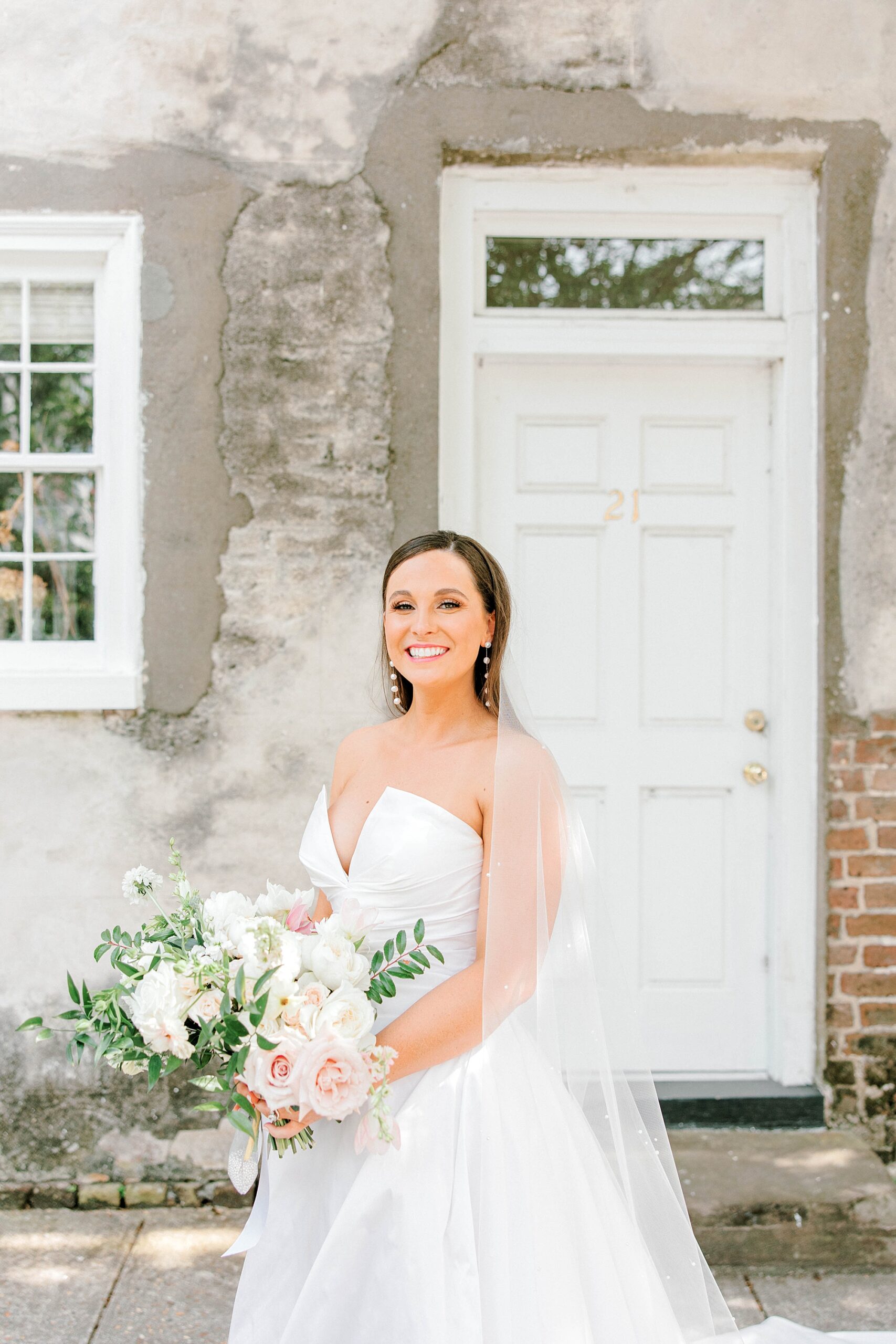brunette bride holds bouquet of ivory and pink flowers in front of brick homes in Charleston SC