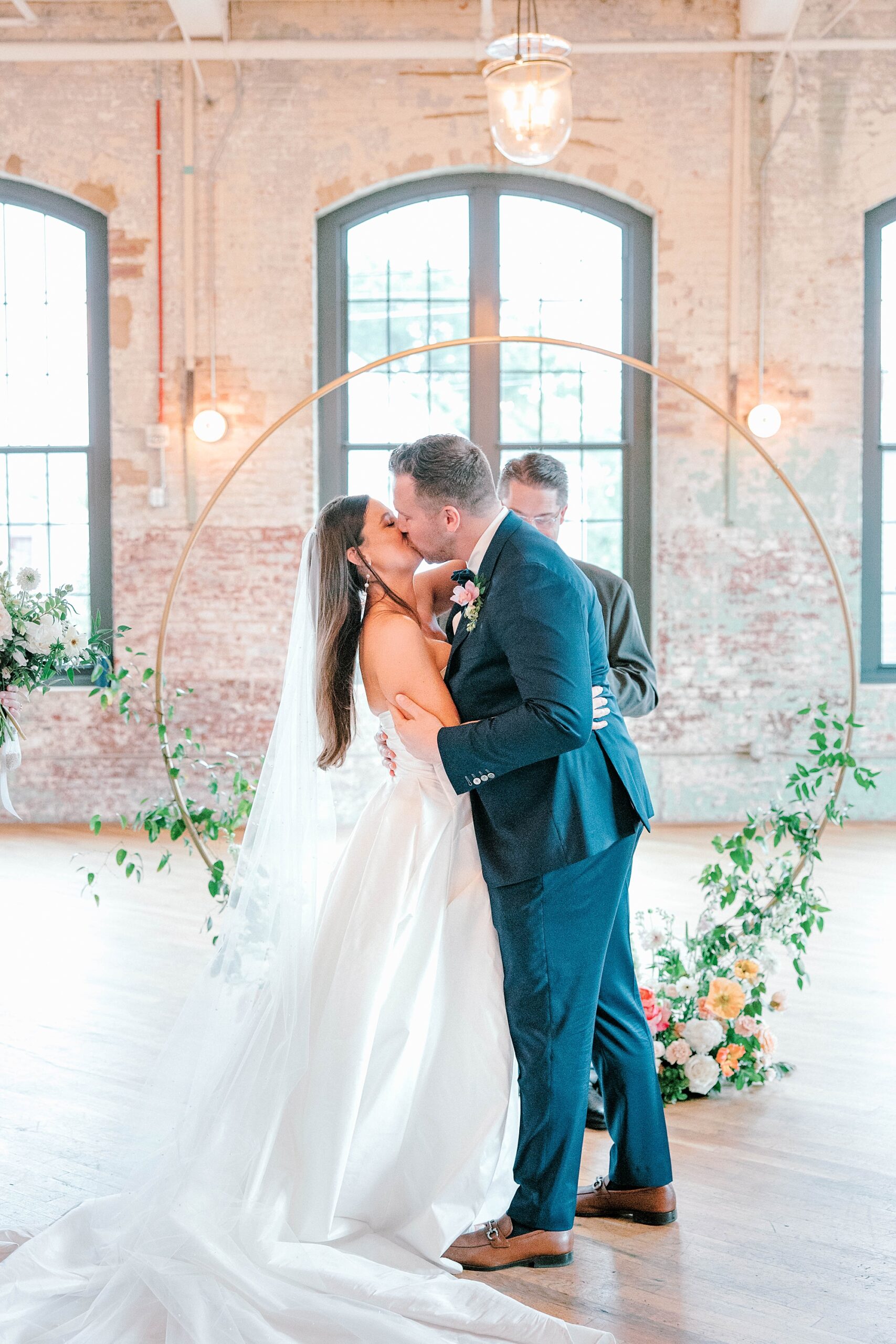newlyweds kiss in front of gold arbor with pink and peach roses inside The Cedar Room