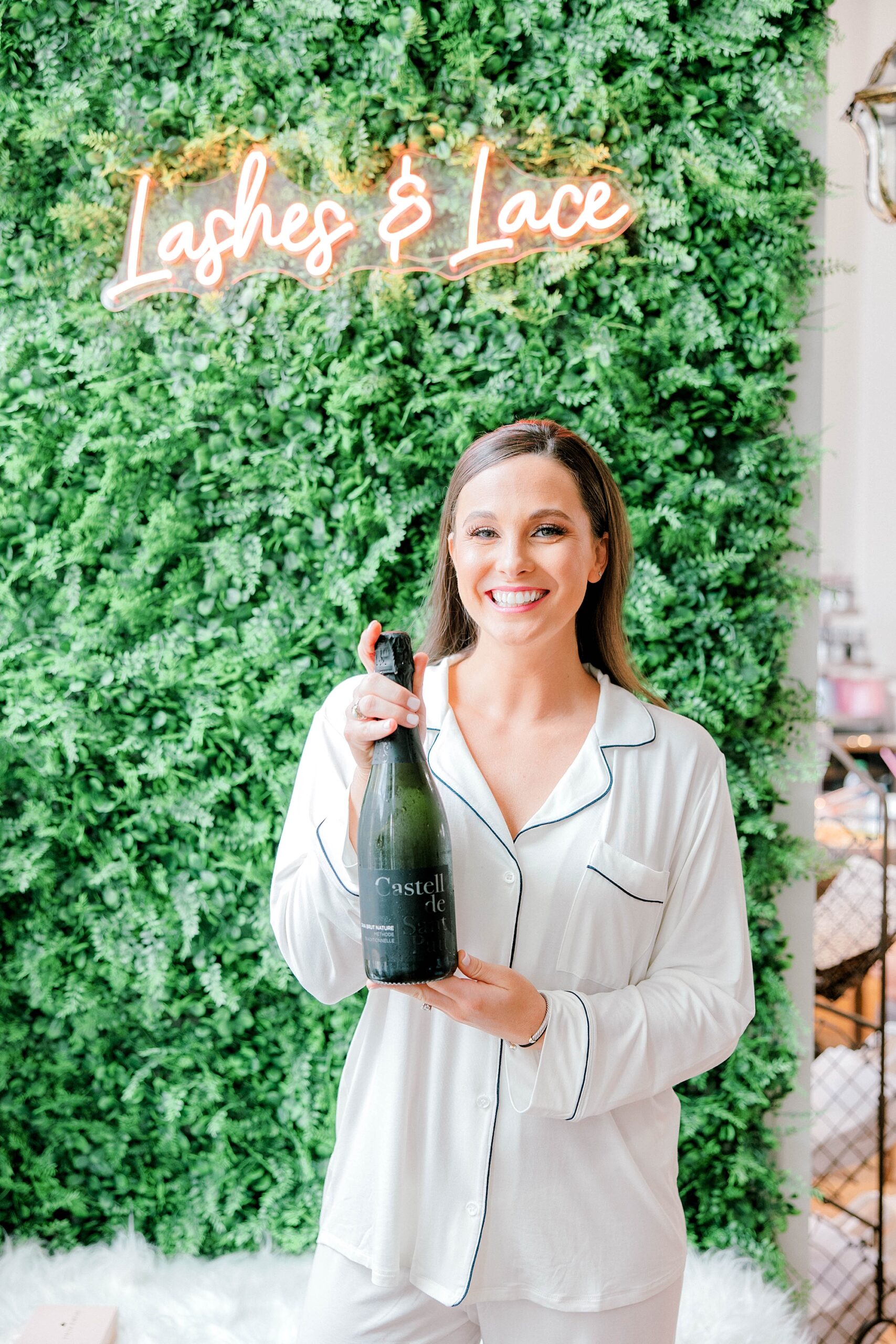 bride holds bottle of champagne in white pajamas by greenery wall with neon sign at Lashes & Lace