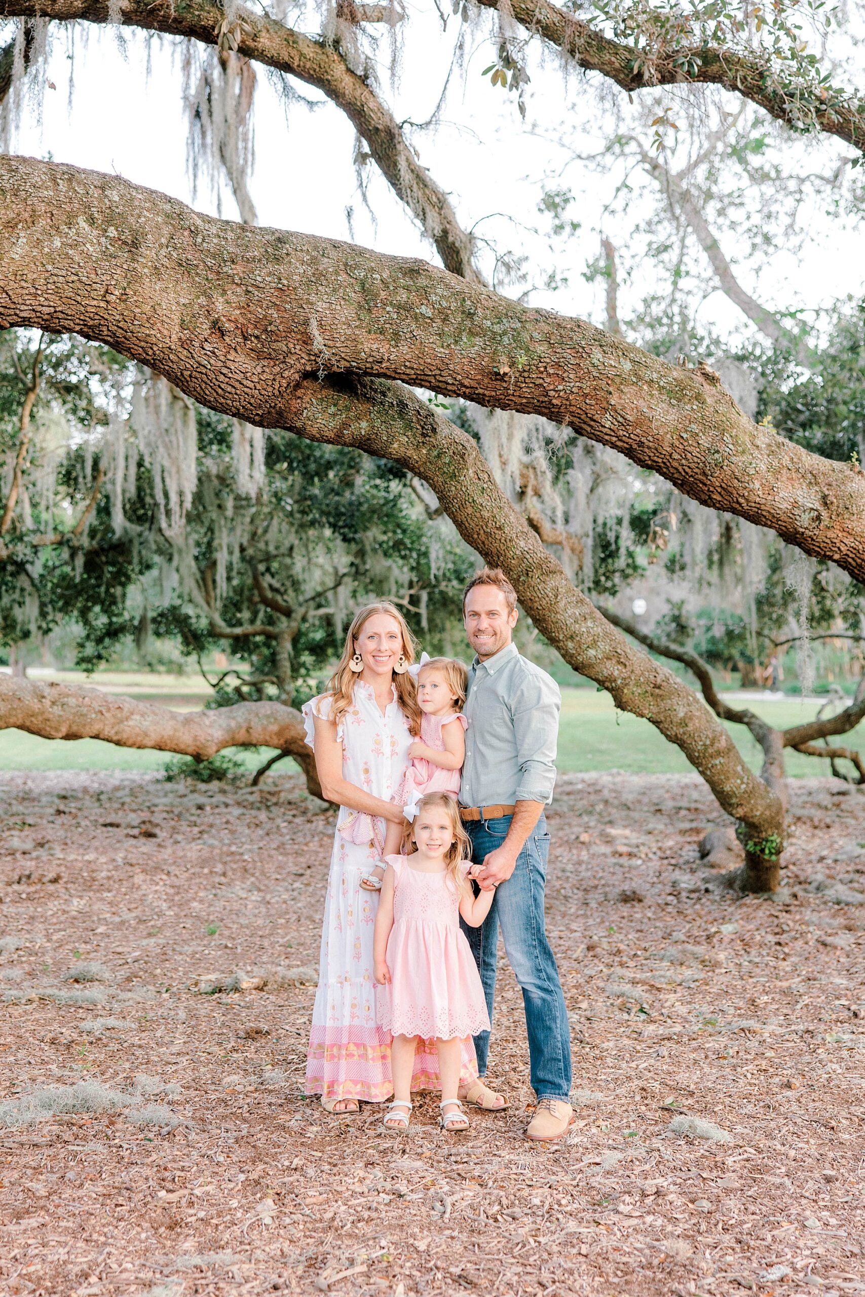 married couple stands with two daughters in pink dresses under tree in Charleston park