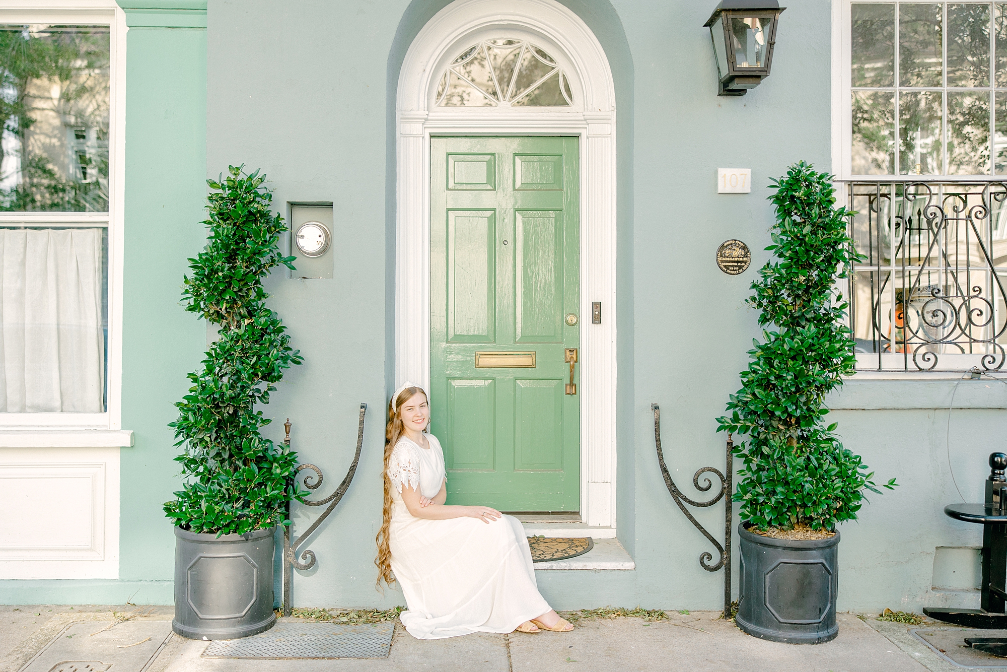 senior in white dress sits by blue and green house on Rainbow Row in Charleston SC