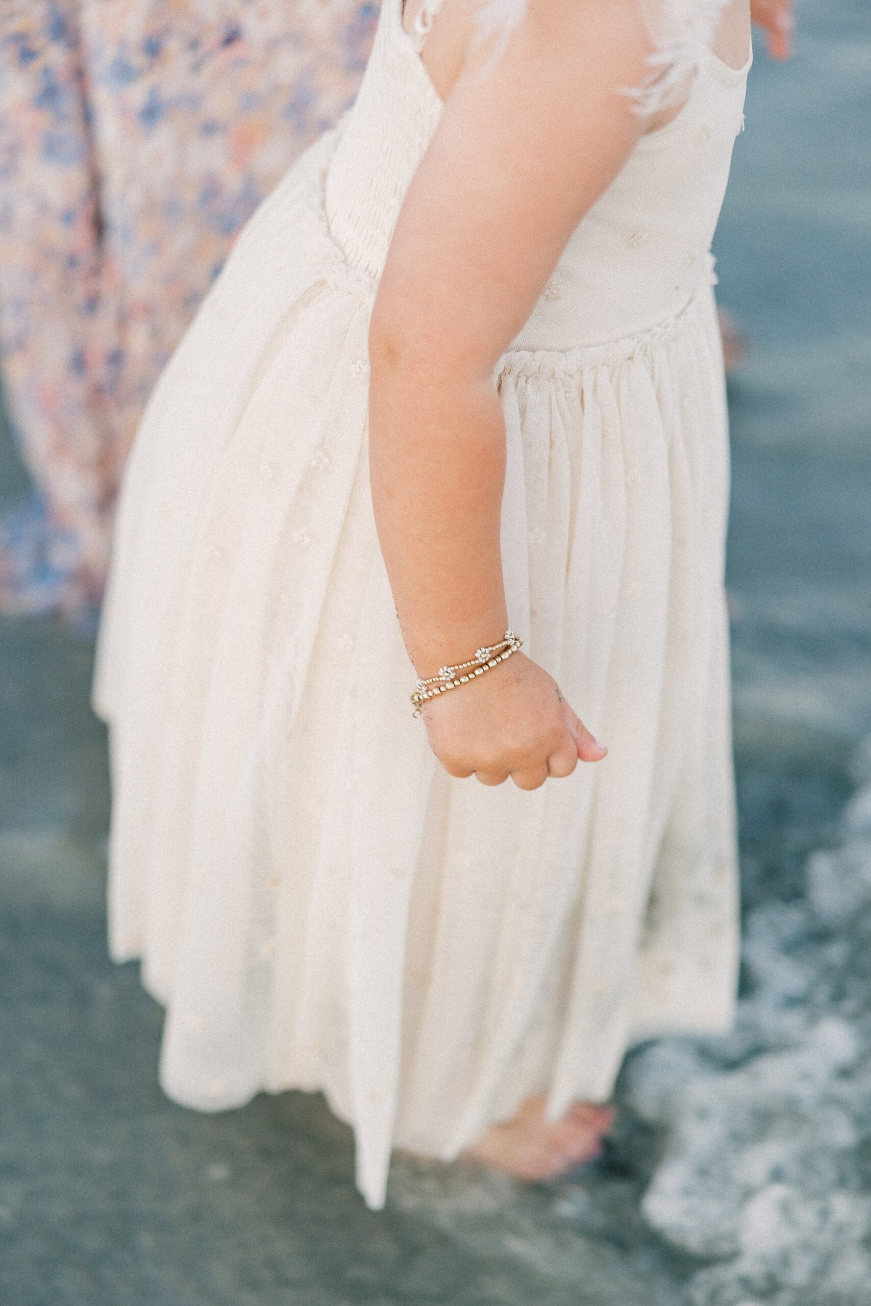 young girl stands with waves crashing over feet showing off bracelet on her arm
