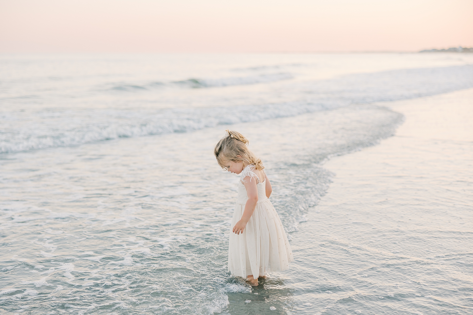 toddler girl in white dress stands in waves at high tide during Folly Beach family portraits