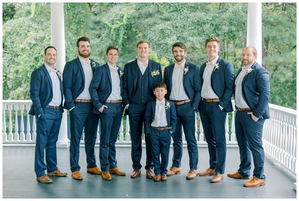 Groom and groomsmen in navy suits and blush bow ties on plantation porch at Old Wide Awake summer wedding in Charleston.